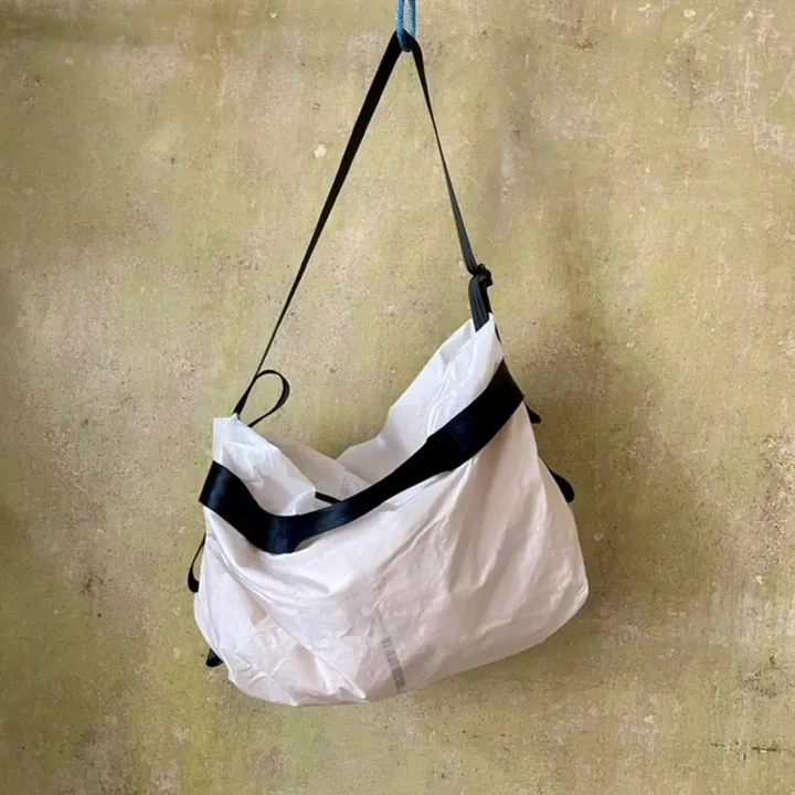 Bílá taška z airbagové tkaniny s černými popruhy, zavěšená na olivové omítnuté stěně.